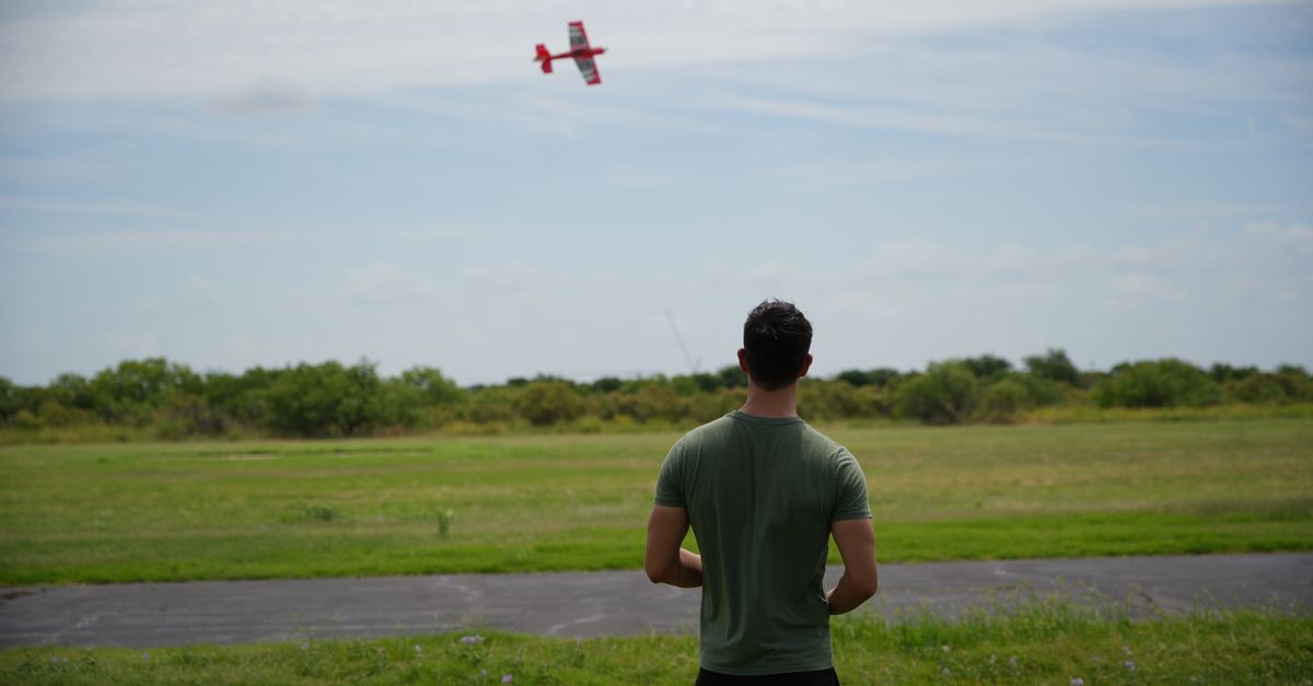 Man flying RC airplane in a green field under a clear sky.